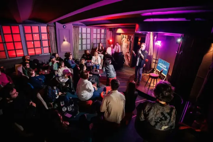 Dimly lit venue with red-backlit windows, host addressing seated audience during a Playing Talking session